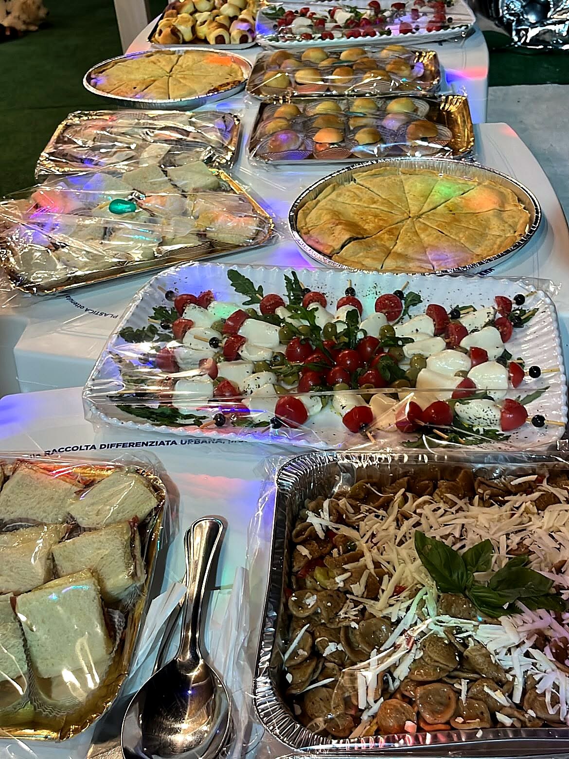 Buffet con vassoi di spiedini caprese, orecchiette al formaggio, tramezzini e torte salate coperti da pellicola